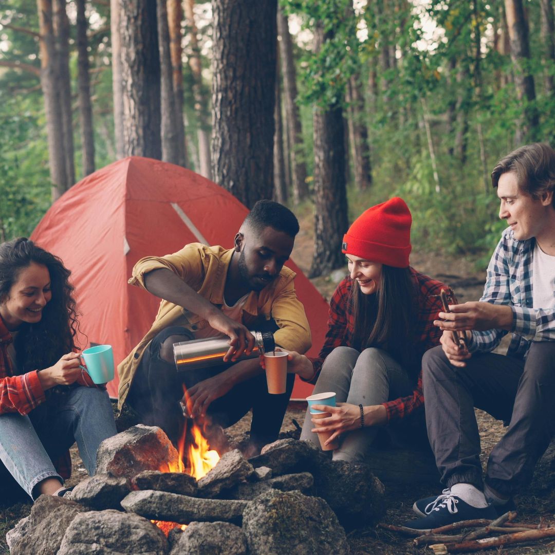 Group of friends sitting around a campfire in the woods with a tent in the background.