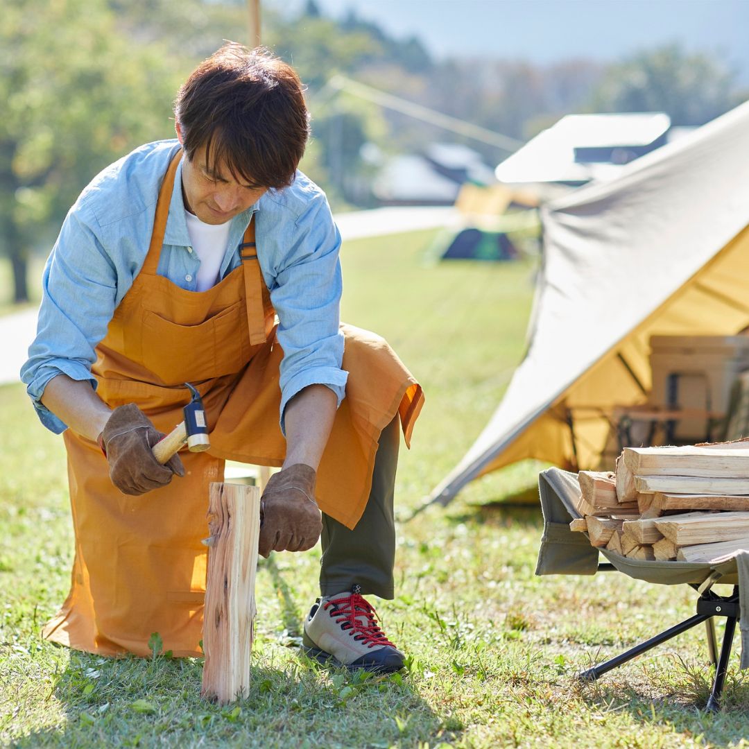 Person in an orange apron chopping wood outdoors with a tent and camping gear in the background.