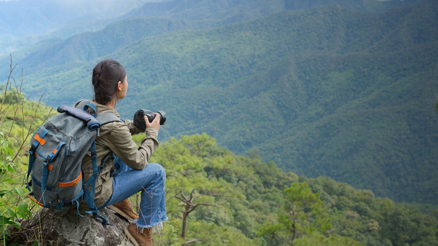 Woman with backpack taking photo of vast green mountain landscape from rocky peak